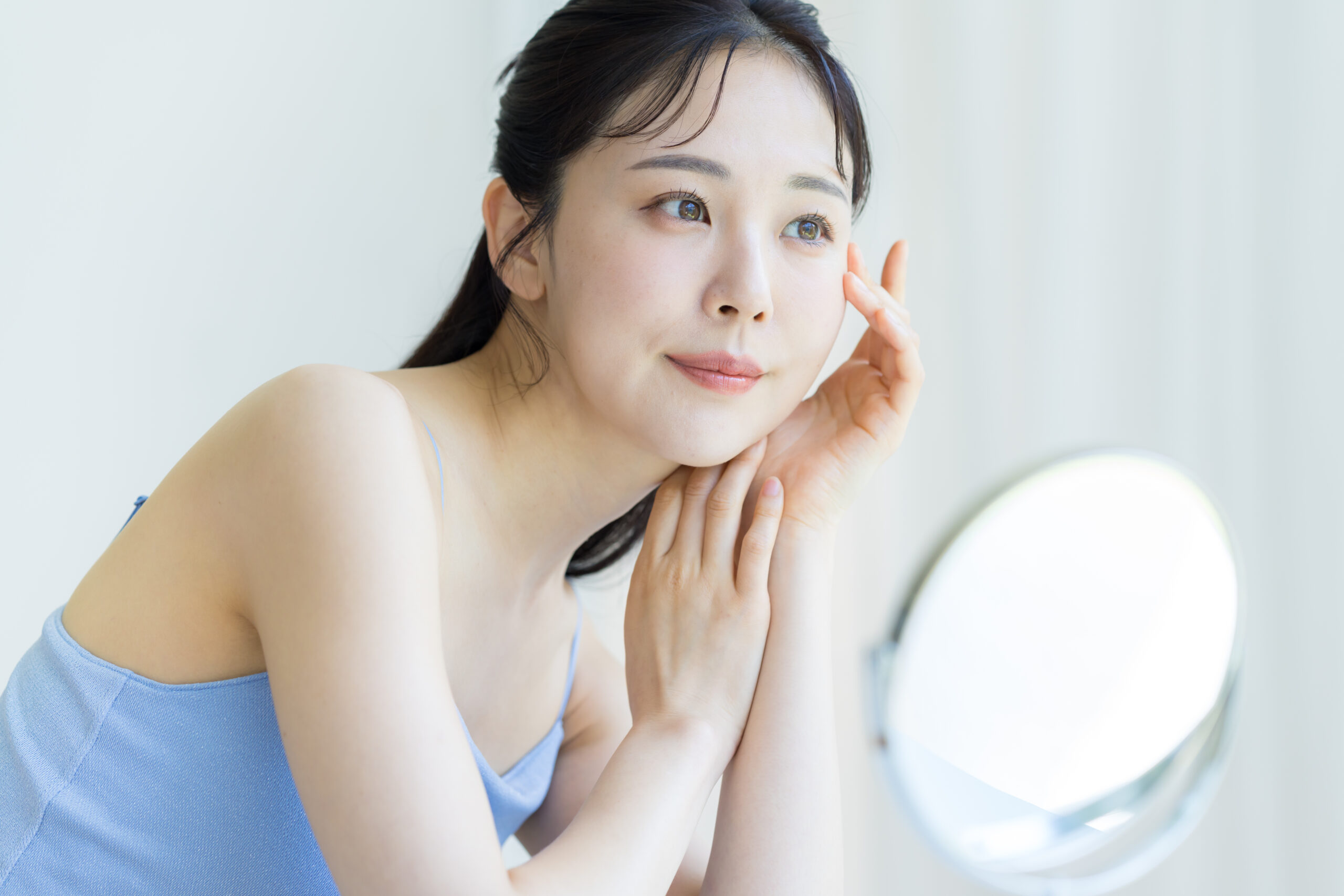 光の入る窓辺で鏡を見ながら肌を確かめる女性